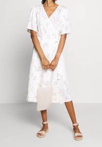 Robe portefeuille blanche à motif floral, manches courtes et encolure en V. Comprend un sac texturé de couleur claire et des sandales beiges.