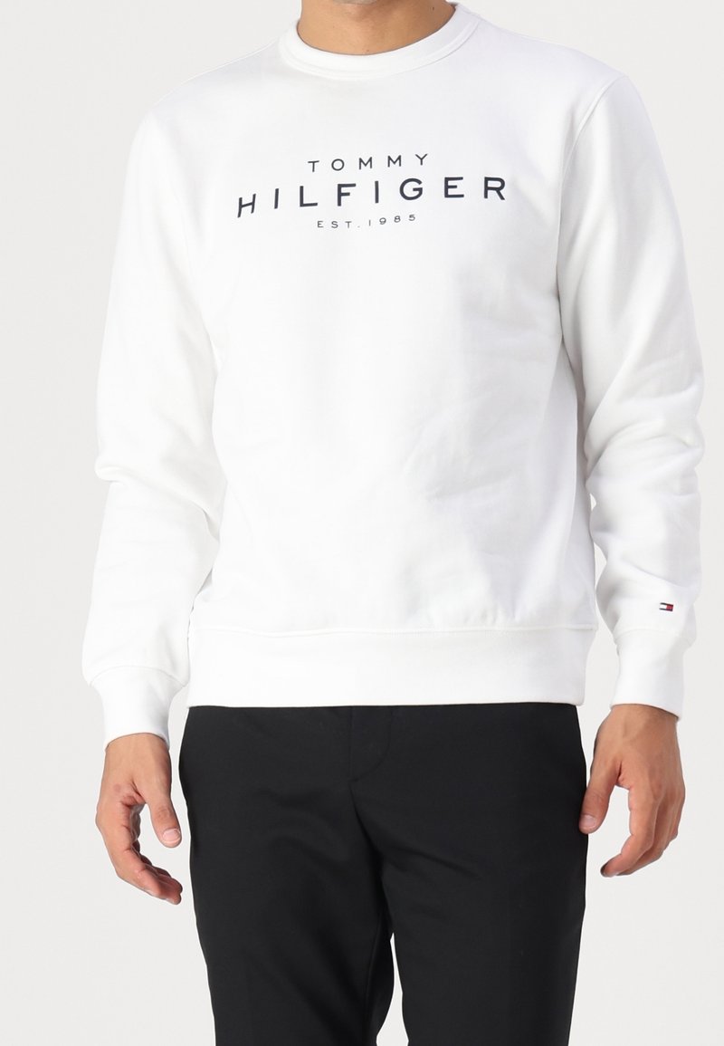 Homme portant un sweat-shirt blanc à manches longues avec le texte "Tommy Hilfiger EST. 1985" et un petit logo sur la manche gauche, associé à un pantalon noir.