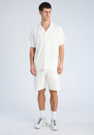 Jonge man staat, draagt een wit shirt met korte mouwen en bijpassende korte broek, witte sokken en witte sneakers met meerkleurige veters.