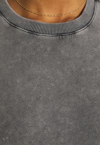 Ingrandimento di una persona che indossa una sottile collana d'oro e un maglione grigio a coste con scollo rotondo.