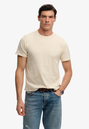 Mand iført beige kortærmet t-shirt, blå jeans med brunt bælte og armbåndsur, stående med den ene hånd i lommen mod hvid baggrund.