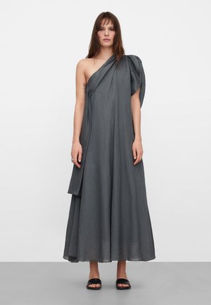 Kobieta ubrana w długą, asymetryczną suknię w ciemnoszarym kolorze z drapowanym materiałem oraz czarne, otwarte sandały na płaskiej podeszwie, stojąca na jednolitym tle.