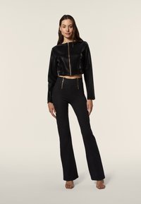 Giubbotto cropped in finta pelle nera con zip frontale e maniche lunghe, abbinato a pantaloni a vita alta neri svasati con dettagli in zip dorati.
