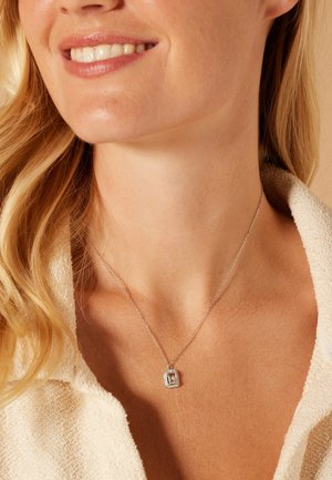 Collier en argent avec pendentif rectangulaire, comportant une pierre centrale entourée d'une bordure texturée, porté sur un tissu de couleur claire.