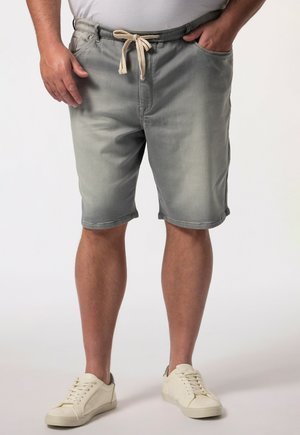 Mand iført grå shorts med snøre og hvide sneakers står med den ene hånd i lommen foran en ensartet baggrund.