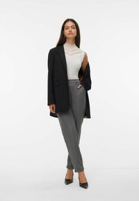 Mujer con un blazer negro caído sobre los hombros, top sin mangas de encaje blanco con cuello alto, pantalones grises de rayas, y tacones negros de punta.