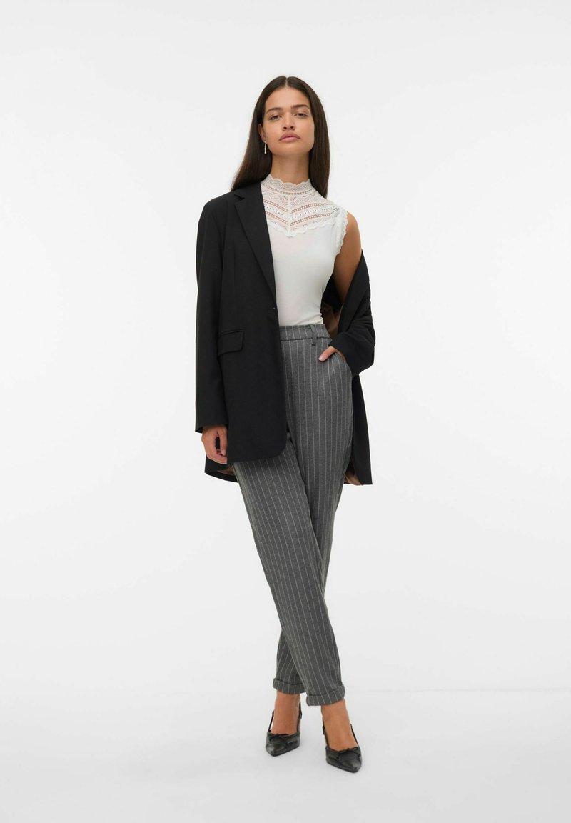Mujer con un blazer negro caído sobre los hombros, top sin mangas de encaje blanco con cuello alto, pantalones grises de rayas, y tacones negros de punta.