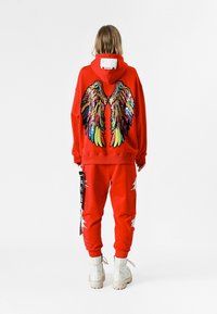 Sudadera roja con un diseño de alas coloridas y con lentejuelas en la parte posterior, combinada con pantalones rojos a juego que presentan acentos blancos y botas gruesas de color crema.