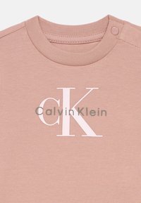 Lys rosa bomull T-skjorte med rund hals, med en hvit logo "CalvinKlein" og et fremtredende "cK" i rosa. Inkluderer trykknapper på skuldrene.