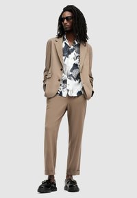 Chaqueta y pantalones de traje beige con corte ajustado. Camisa de patrón en blanco y negro por debajo, complementada con gafas de sol negras y zapatos.