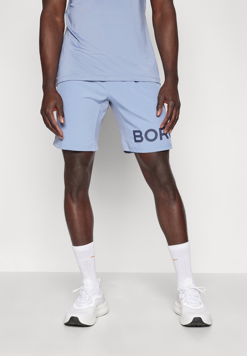 Calções de desporto azul claro com uma textura em malha, apresentando um grande gráfico em azul escuro com a palavra "BORN" na perna esquerda, acompanhados de ténis brancos.