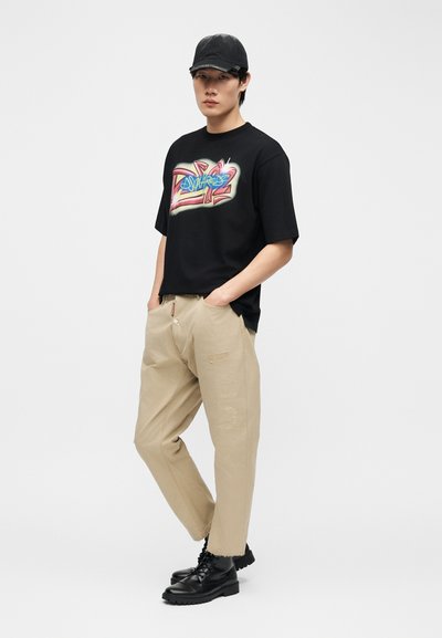 Jeune personne portant un t-shirt graphique noir style graffiti, pantalon beige avec détails en patch, casquette noire et bottes noires à lacets, debout les mains dans les poches.