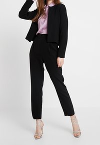 Femme portant un pantalon noir cintré, une veste courte noire, un chemisier violet clair et des sandales argentées à talons hauts à brides, sur fond blanc.