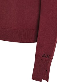 Manica di un maglione a maglia bordeaux con triangolo ricamato in nero e simboli a X vicino al polsino, con una piccola fessura in tessuto sul bordo del polsino.