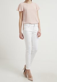 Světle růžová blůza s krátkými volánkovými rukávy, kombinovaná s bílými skinny džíny. Model má na sobě nahé vysoké podpatky. Jednoduché a čisté linie po celém outfitu.