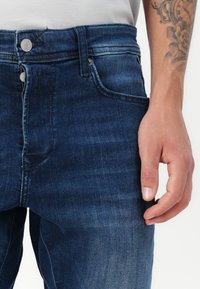 Tummansiniset farkut, joissa on klassinen viiden taskun muotoilu, valmistettu hieman karheasta denimmateriaalista. Kuvassa on käsi lepäämässä lähellä.