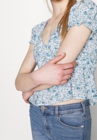 Blusa floreale azzurro chiaro con maniche corte a palloncino, scollo a v e orlo peplum, abbinata a jeans in denim blu. Presenta piccoli fiori e delicate rifiniture a chiocciola.