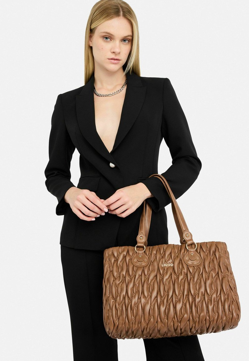 Geantă de tote din piele maro matlasată, cu mânere duble, având cusături decorative și accente din metal, purtată de o persoană îmbrăcată într-un costum negru.