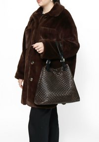 Geweven bruine leren handtas met een gestructureerd patroon, zwarte handvaten en een slouchy vorm, vastgehouden door een persoon in een donkerbruine jas.