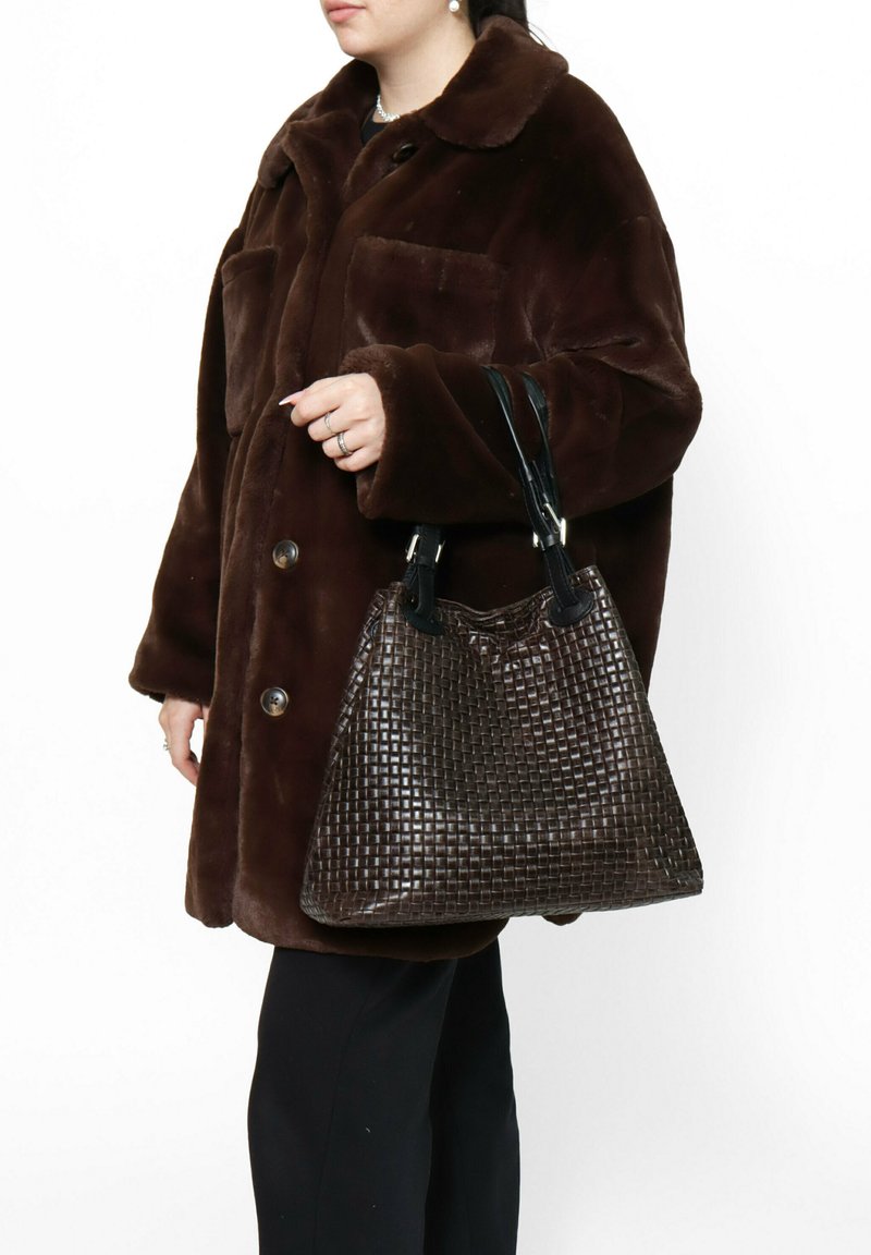 Geweven bruine leren handtas met een gestructureerd patroon, zwarte handvaten en een slouchy vorm, vastgehouden door een persoon in een donkerbruine jas.