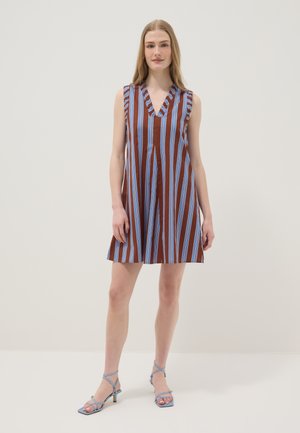 Jeune femme debout, portant une robe marron sans manches avec des rayures bleues verticales et un col en V à volants, associée à des talons à brides bleu clair.