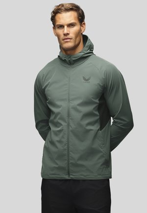 Homme aux cheveux châtain clair portant une veste à capuche zippée verte et un pantalon noir, debout les mains dans le dos sur un fond gris.
