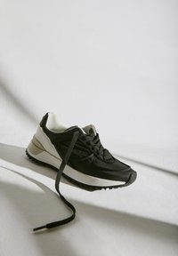 Zapatilla deportiva negra con parte superior texturizada y acolchada, media suela blanca en contraste y suela exterior de goma negra. Presenta puntera redonda y cordones.