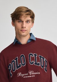 Ung man med ljusbrunt hår bär en vinröd sweatshirt med texten "POLO CLUB Beyond Classics Established 2012" över en blå skjorta.