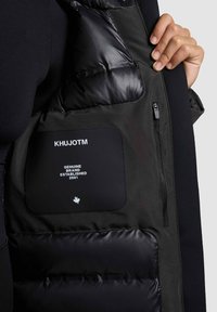Čierna prešívaná vesta KHUJOTM s hladkým, lesklým materiálom, so zipsovým vreckom a matným nášivkom s logom a textom značky.