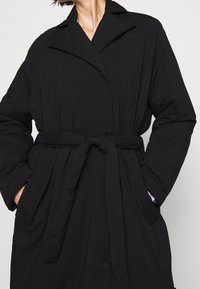 Schwarze gepolsterte Jacke mit Kragen, langen Ärmeln und Bindegürtel. Sie verfügt über Seitentaschen und einen strukturierten Stoff.