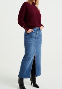 Denimová maxi sukně s předním rozparkem, v kombinaci s burgundským pleteným svetr, který má zlaté knoflíkové detaily na rukávech. Černé vysoké boty na podpatku.