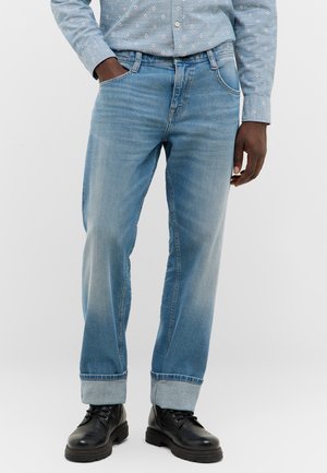 Man wearing light blue cuffed jeans, a patterned blue shirt with one hand in pocket, and black lace-up boots on a white background.