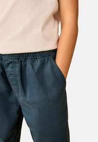 Pantalon en coton bleu foncé avec une taille élastique et des poches sur les côtés, associé à une chemise à manches courtes beige clair. Texture lisse.