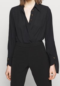 Blusa nera avvolgente con colletto, dotata di bottoni in accento dorato e maniche lunghe con polsini abbottonati. Tessuto liscio con un design leggermente drappeggiato.