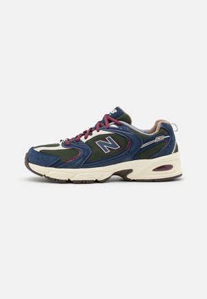Chaussure de sport pour homme avec mesh vert, superpositions en daim bleu marine, lacets bordeaux et semelle blanche avec détail de coussinage visible.