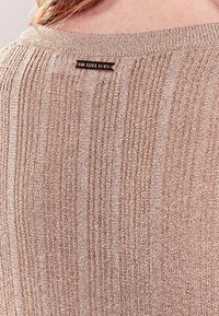 Primer plano de un suéter de punto acanalado beige con la placa de metal del logo de Michael Kors cerca del cuello, en la espalda de una persona.