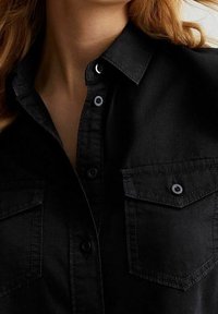 Donna che indossa una camicia nera con bottoni, tasche frontali e colletto, che mostra parte del suo collo e i capelli mossi di colore castano chiaro.