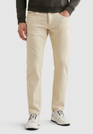 Béžové slim-fit rifle nosené s bielymi teniskami a tmavozeleným mikinou, ruky voľne pri tele.