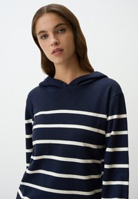 Felpa blu navy con strisce orizzontali bianche, tessuto morbido, maniche lunghe e vestibilità rilassata. Presenta un cappuccio con coulisse.