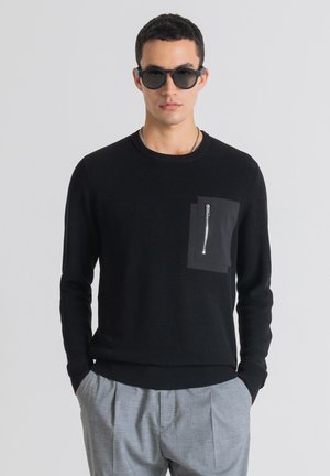 Maglione nero a coste con tasca frontale con zip, abbinato a pantaloni grigi a pieghe. Il modello indossa occhiali da sole circolari scuri.