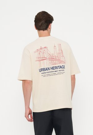 Άνδρας που φορά ελαφρύ T-shirt με κόκκινο σκίτσο της Γέφυρας Μπρούκλιν και του ορίζοντα της Νέας Υόρκης και κείμενο "Urban Heritage" στην πλάτη.