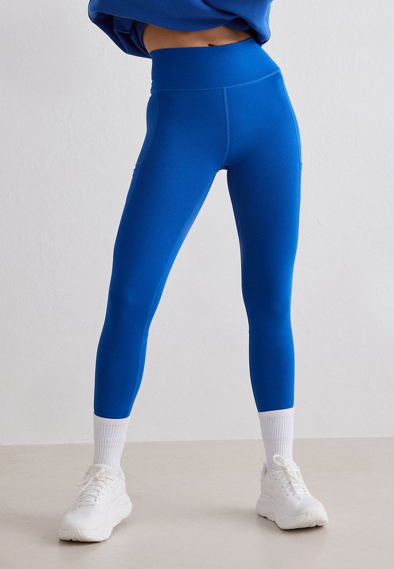 Jambes vêtues de leggings taille haute bleu vif, de chaussettes blanches mi-mollet et de baskets blanches épaisses, sur un fond clair uni.