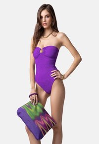 Costume da bagno viola senza spalline con trama a coste e anello decorativo al centro. Pochette stampata verde e viola in mano, con un motivo a onde.