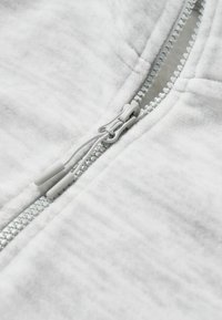 šedá mikina s kapucí na zip, která se pyšní měkkou fleecovou tkaninou, stříbrným zipem se stahovacími pásky a plochými švy. Detailní pohled ukazuje texturu a šití.