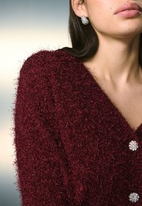 Vinrød, struktureret sweater med blødt garn, der har en dyb V-hals og glitrende runde knapper. Pyntet med juvelbesatte øreringe.