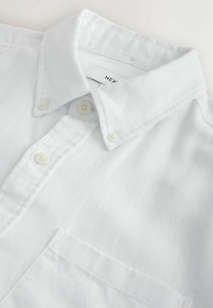 Cămașă albă cu nasturi, guler structurat, textura țesăturii este netedă, cu un buzunar pe piept și nasturi mici pe față.