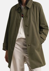 Zelený kabát na knoflíky přes bílý svetr s copánkovým vzorem a světle béžové kalhoty, s jednou rukou v kapse kabátu.