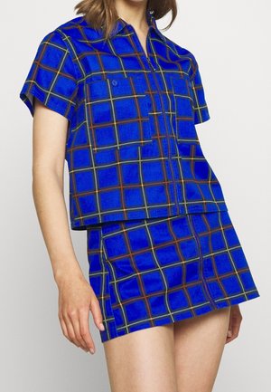 Kvinde iført blå ternet kortærmet skjorte og matchende miniskørt med frontlynlås mod hvid baggrund.