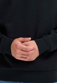 Sudadera negra de manga larga, con puños y dobladillo acanalados. Manos entrelazadas al frente, mostrando el tono de piel natural y detalles de las uñas.