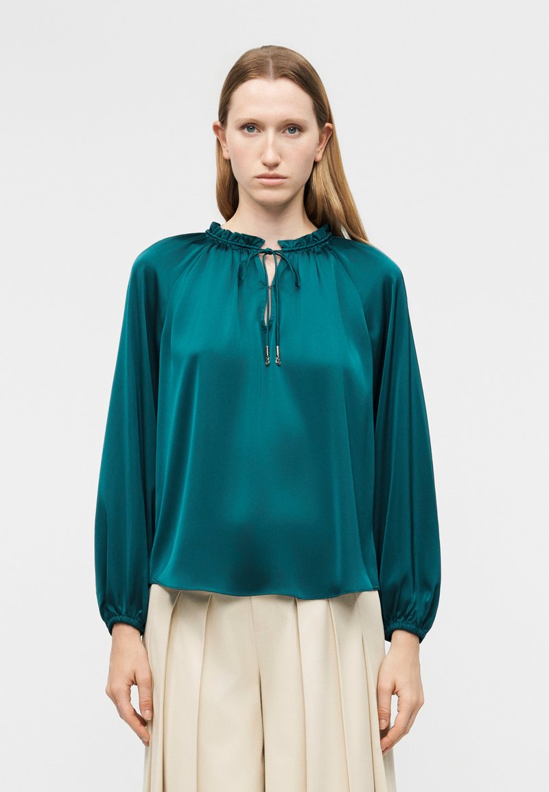 Γυναίκα που φοράει μπλούζα από σατέν σε τιρκουάζ με μακριά μανίκια, φραμπαλά στο λαιμό και κορδόνι, συνδυασμένη με μπεζ πλισέ παντελόνι.